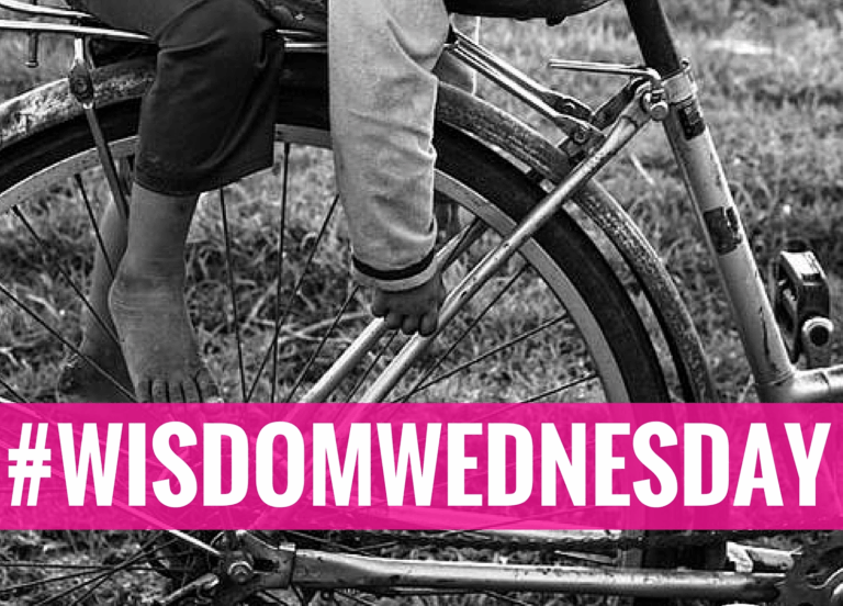 Black-and-white photo of a child sitting on a bicycle, with bare feet on the pedals. A bold pink banner with white text reads #WISDOMWEDNESDAY across the bottom of the image.