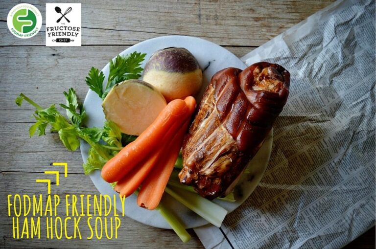 A plate with a smoked ham hock, carrots, celery, celery leaves, and a swede on a marble board, beside newspaper. Logos indicate FODMAP Friendly and Fructose Friendly. Text reads: FODMAP Friendly Ham Hock Soup.