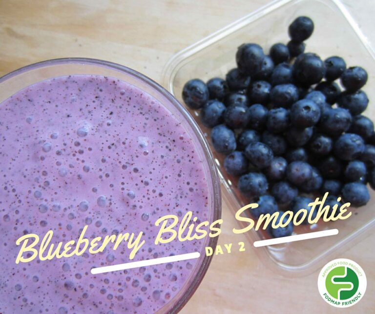A glass of purple blueberry smoothie sits next to a punnet of fresh blueberries on a light wood surface. Text reads “Blueberry Bliss Smoothie Day 2” with a green FODMAP Friendly logo in the corner.