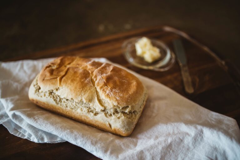 A loaf of rustic bread rests on a white cloth atop a wooden surface, with a butter knife and a small dish of butter placed in the background.