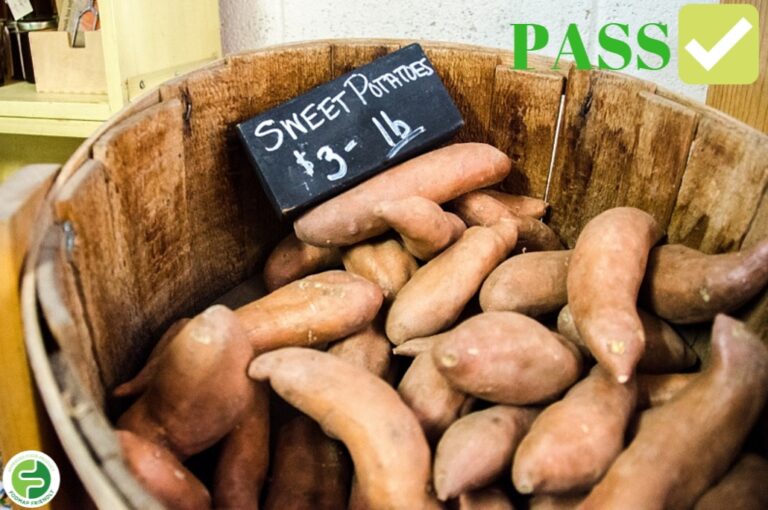 A wooden basket filled with sweet potatoes sits under a chalkboard sign that reads Sweet Potatoes £3 - 1 lb. A green PASS symbol with a tick is displayed in the top right corner.