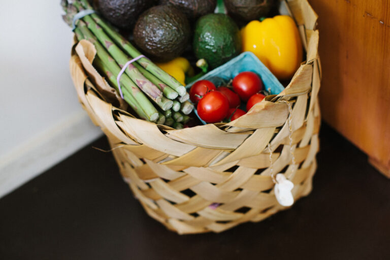 A woven basket filled with fresh vegetables, including asparagus, avocados, cherry tomatoes in a blue punnet, and a yellow pepper, sits on a dark floor next to a wooden surface.