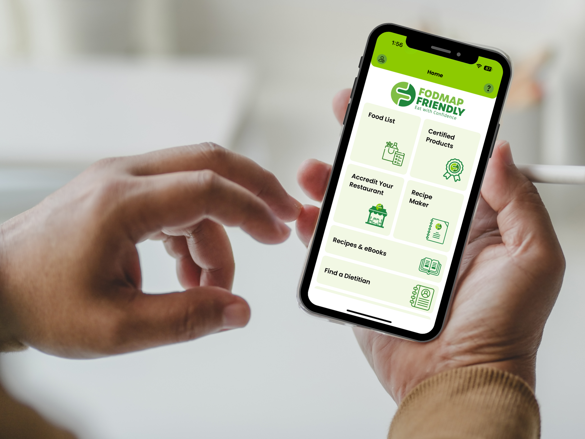 A person holds a smartphone displaying the FODMAP Friendly app, featuring menu options like Food List, Certified Products, Register, Accredit Your Restaurant, Recipe Master, Recipes & eBooks—all designed to support better gut health.