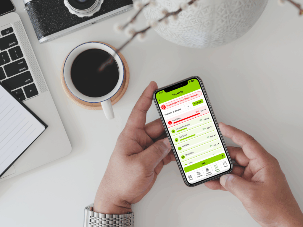 A person holds a mobile phone displaying a gut health app at a desk with a laptop, coffee cup, notepad, and camera, next to a decorative white vase—perfect for Gut Health Professionals looking to register for the latest insights.