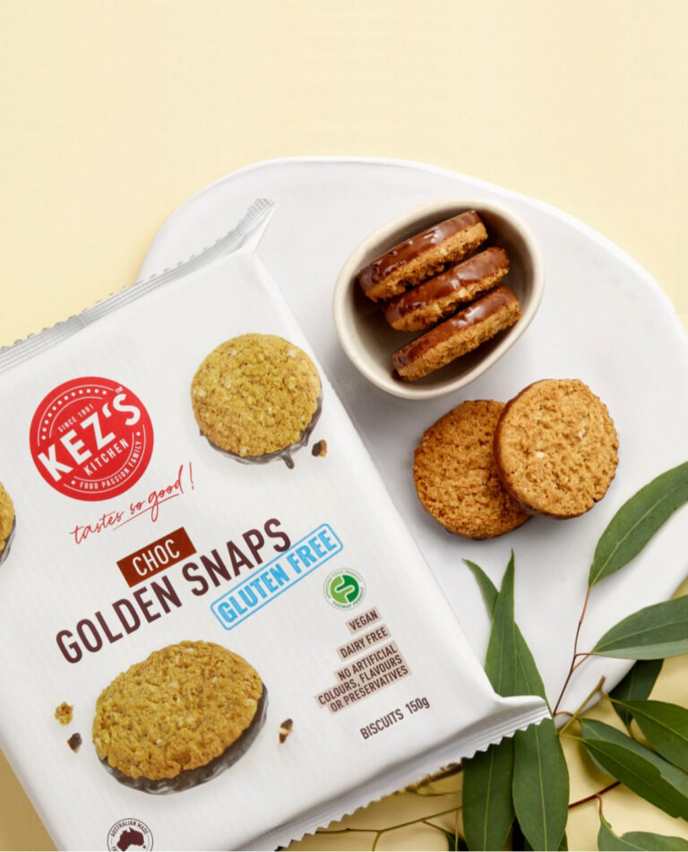 A packet of Kez’s Kitchen Choc Golden Snaps (gluten free, FODMAP Friendly) lies on a white chopping board with some biscuits, two stacked with chocolate filling, next to green leaves on a light yellow background.