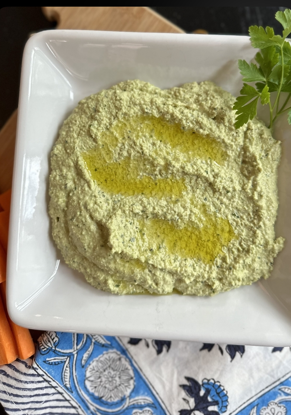 A white dish filled with greenish Low FODMAP houmous drizzled with olive oil, garnished with parsley, and placed on a patterned serviette beside sliced carrots.