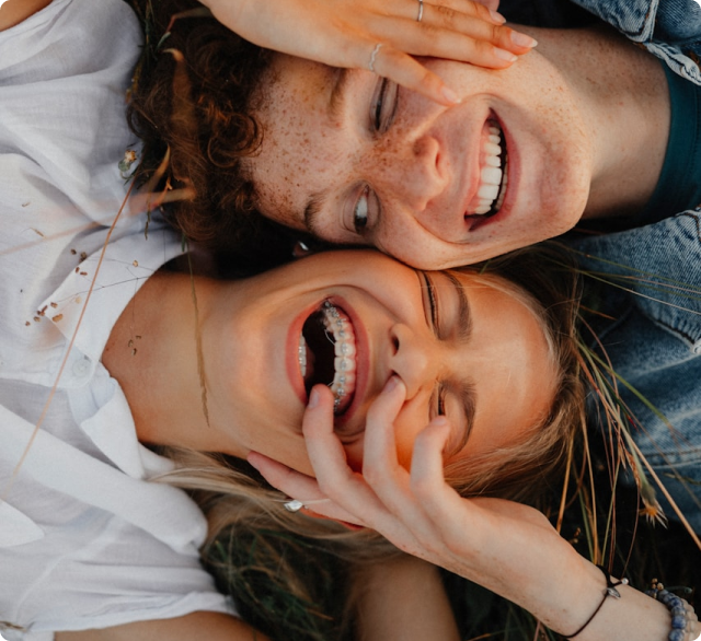 Two smiling people lying on grass, faces close together. One person with braces laughs playfully as the other touches their face, both appearing happy and carefree—showing none of the symptoms that sometimes accompany causes related to a low FODMAP diet.