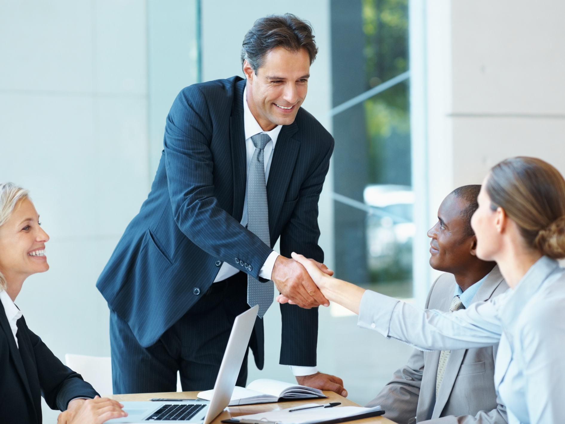 A man in a suit stands and smiles whilst shaking hands with a seated woman in a business meeting, as two other colleagues look on—all appearing engaged and positive during an accreditation discussion.