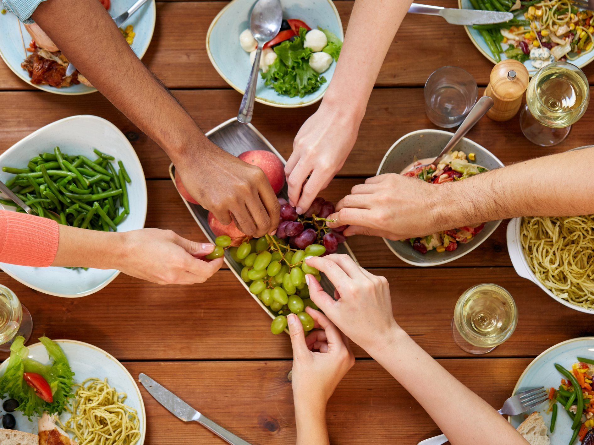 Several hands reach for grapes in a dish at the centre of a wooden table set with Low FODMAP plates of pasta, green beans, salad, and glasses of white wine, creating a communal dining atmosphere perfect for IBS support.