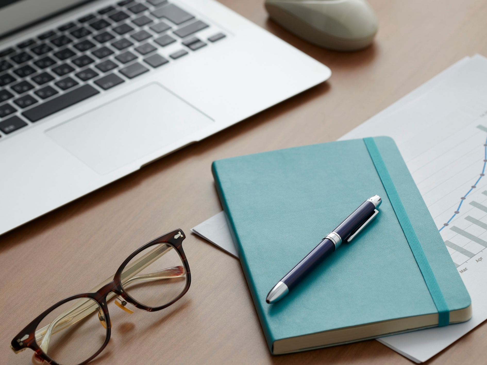 A teal notebook with a pen, spectacles, a laptop, and a graph on paper are arranged on a wooden desk, hinting at professional training or research in gut health within a focused workspace setting.