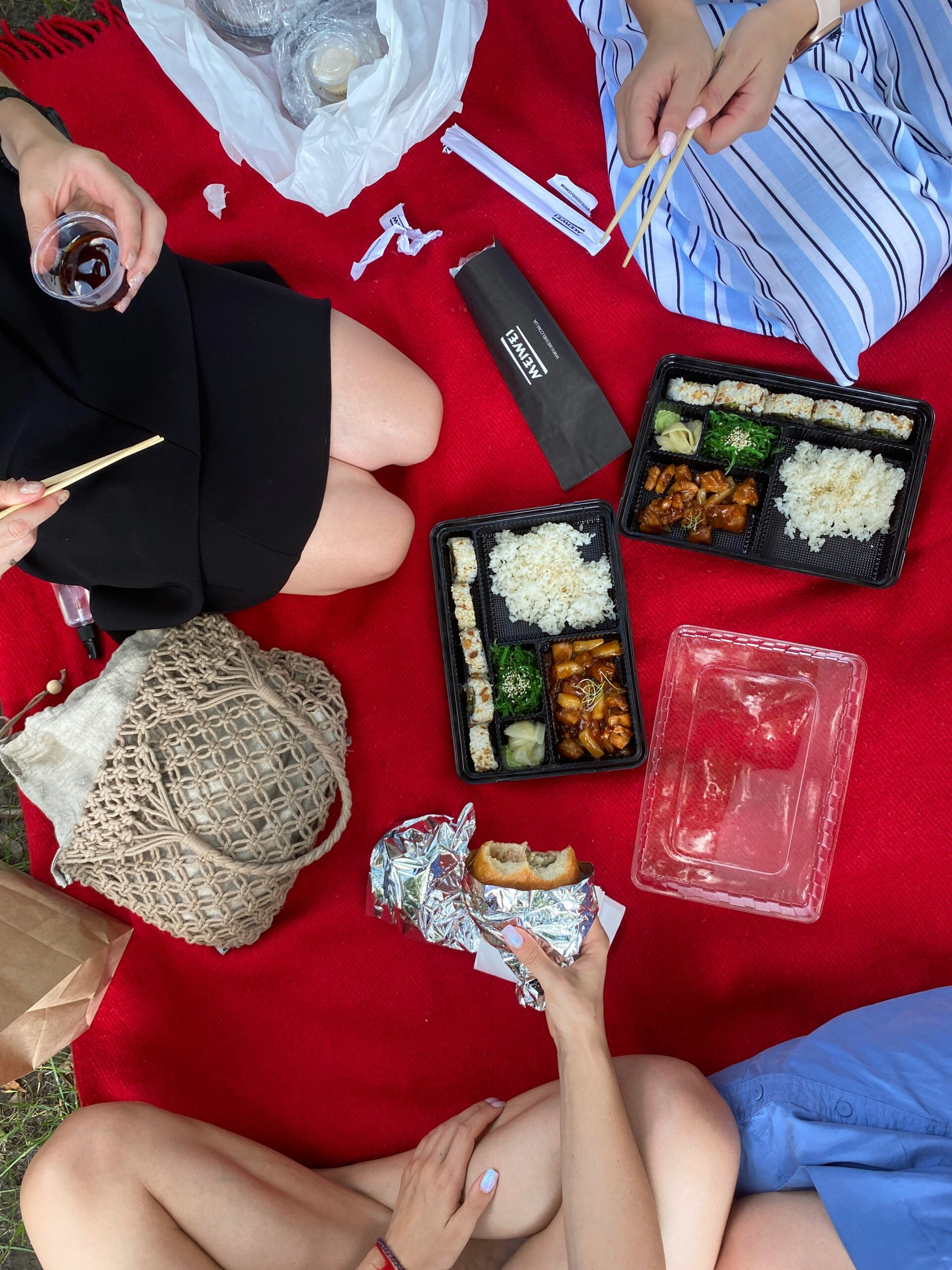 Three people sit on a red picnic blanket, eating takeaway food in black containers with chopsticks—rice, vegetables, dumplings, and meat—enjoying both high FODMAP foods and low FODMAP foods. One holds a sandwich; bags and a drink rest nearby.