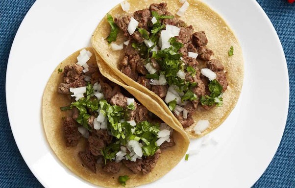 Two corn tortillas filled with chopped grilled beef, diced onions, and fresh coriander, served on a white plate against a blue background.
