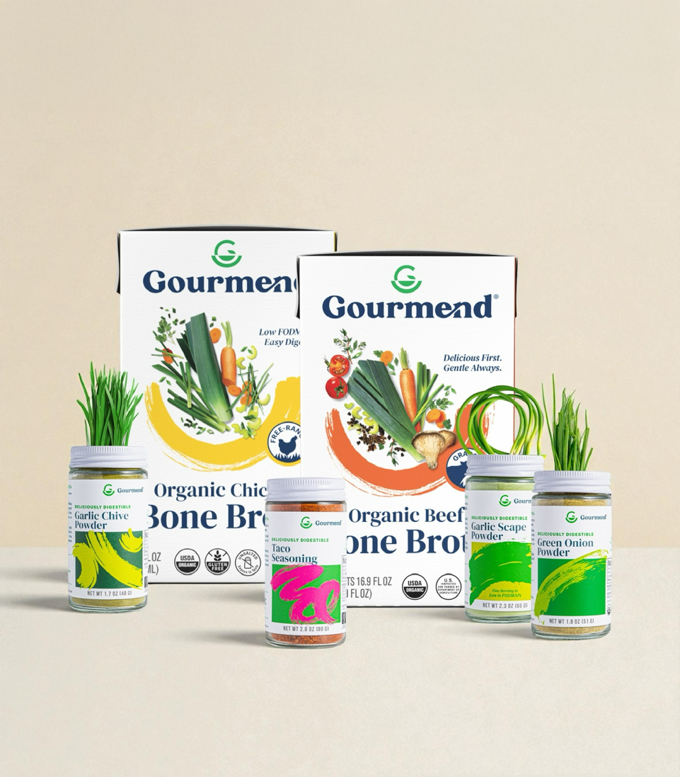 A display of Gourmend products features FODMAP Friendly organic chicken and beef bone broths, plus four jars of seasonings—garlic chive, taco, spring onion, and garlic scape—alongside fresh chives and onions in jars.