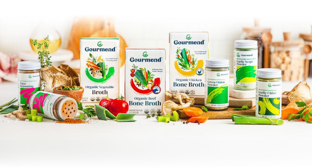 A variety of Gourmend branded, FODMAP Friendly broth and seasoning products are displayed on a white surface with fresh vegetables and herbs, including tomatoes, garlic, and spring onions, arranged around the products.